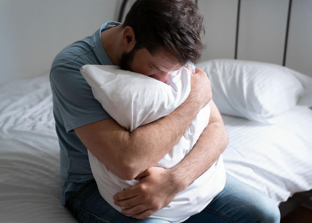 A man suffering from depression sits on his bed and hugs his pillow tightly.