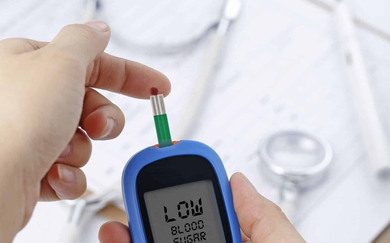 Hand holding a blood glucose meter measuring blood sugar, the background is a stethoscope and chart file